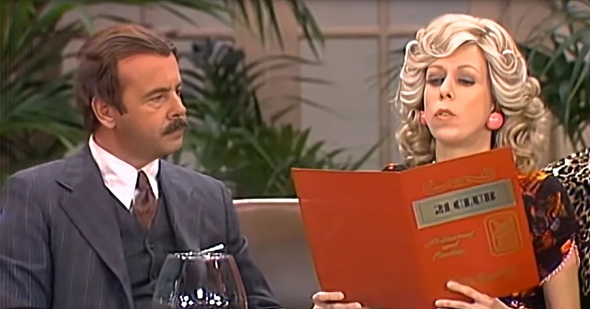 Woman with blonde curls reads book, man in suit looks on, both seated.