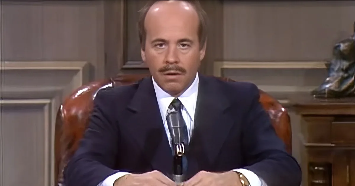 A man in a suit sits at a desk, hands clasped, in a formal setting.