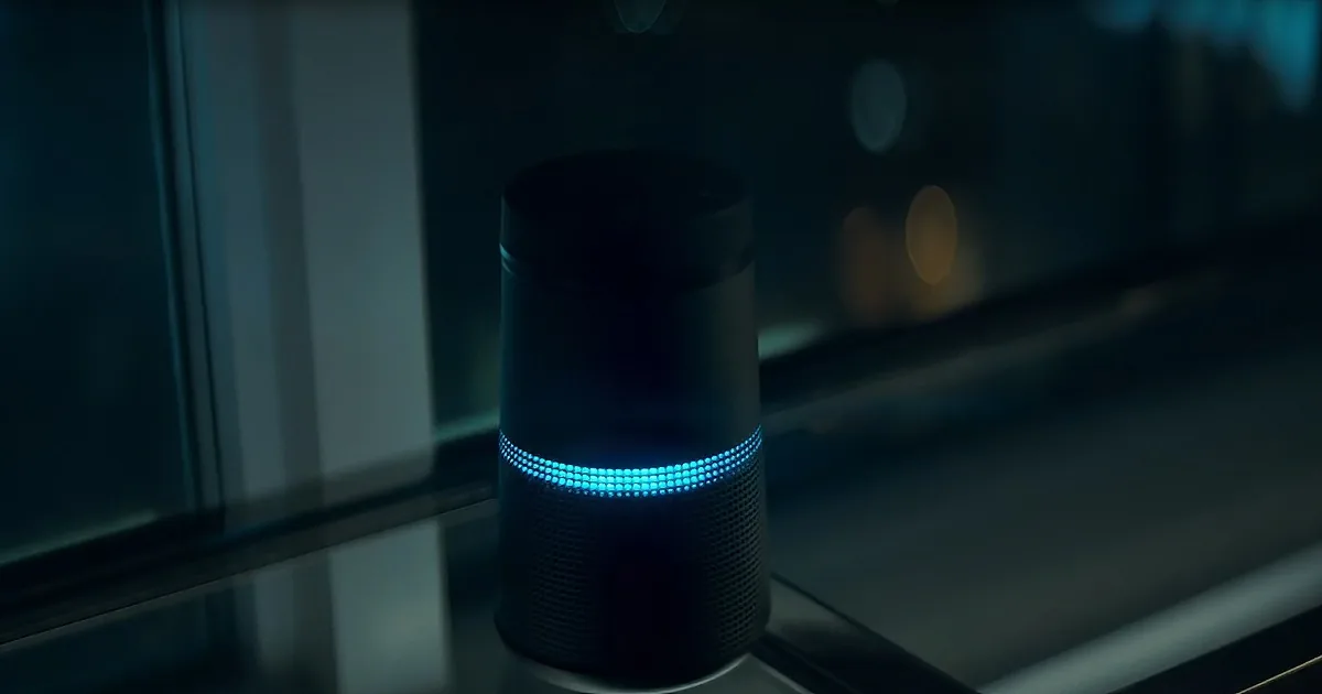 A smart speaker with a glowing blue ring sits on a window sill, illuminated against a dark, blurred background.