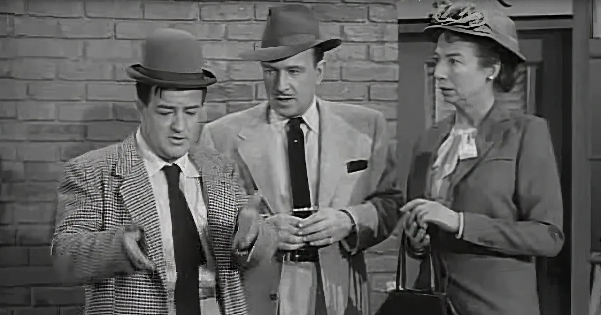 Three individuals in formal attire stand together, one gesturing, in front of a brick wall.