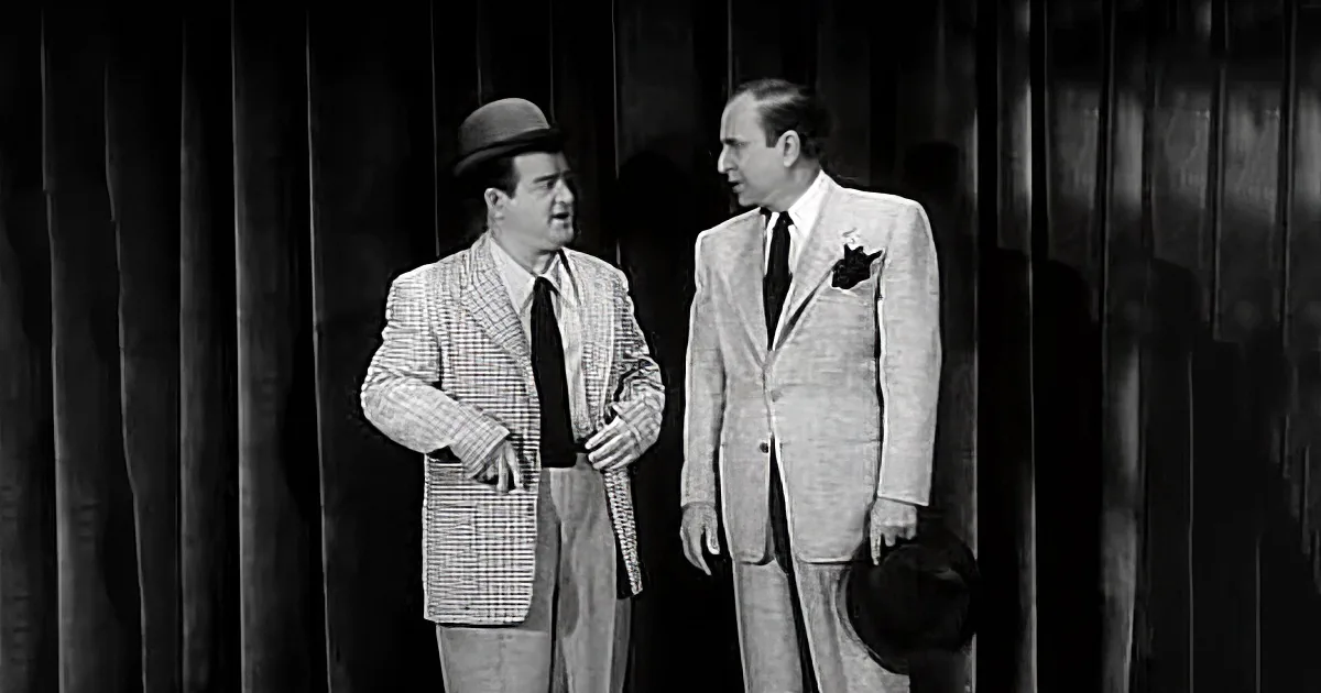 Two men in suits stand against a curtain backdrop, one gesturing with a cane.