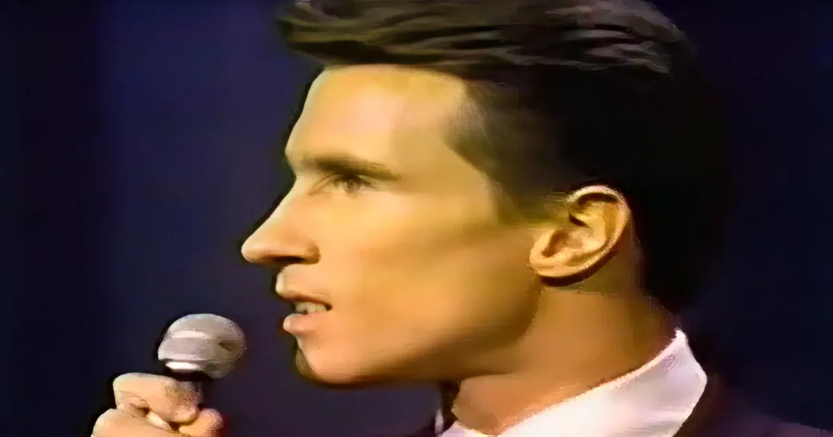 A man with dark hair and a white shirt holds a microphone, singing or speaking.