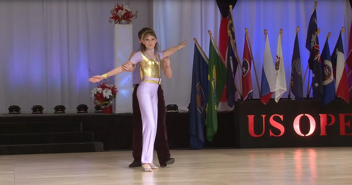 A dancer in a gold and white outfit performs gracefully on stage with flags and "US Open" signage in the background.