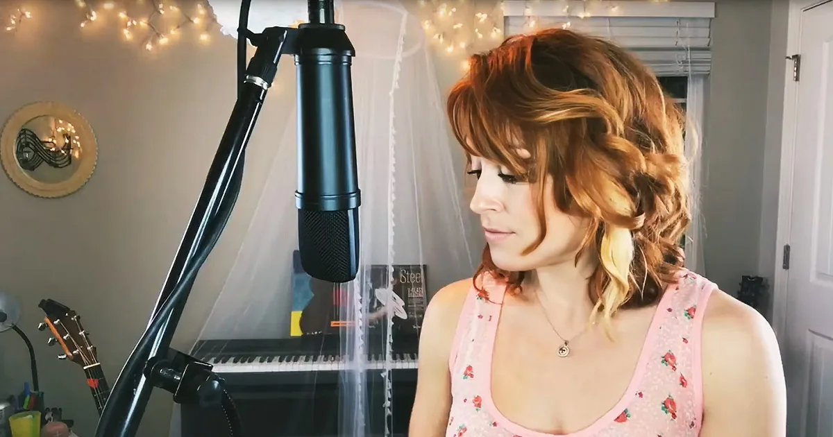 A woman with red hair in a floral top stands in front of a microphone, surrounded by string lights and a piano.