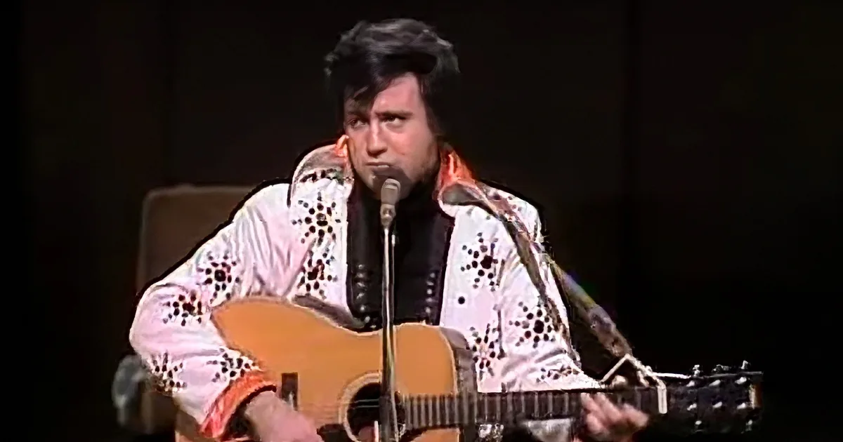 A man in a white jacket with black patterns plays an acoustic guitar and sings into a microphone.