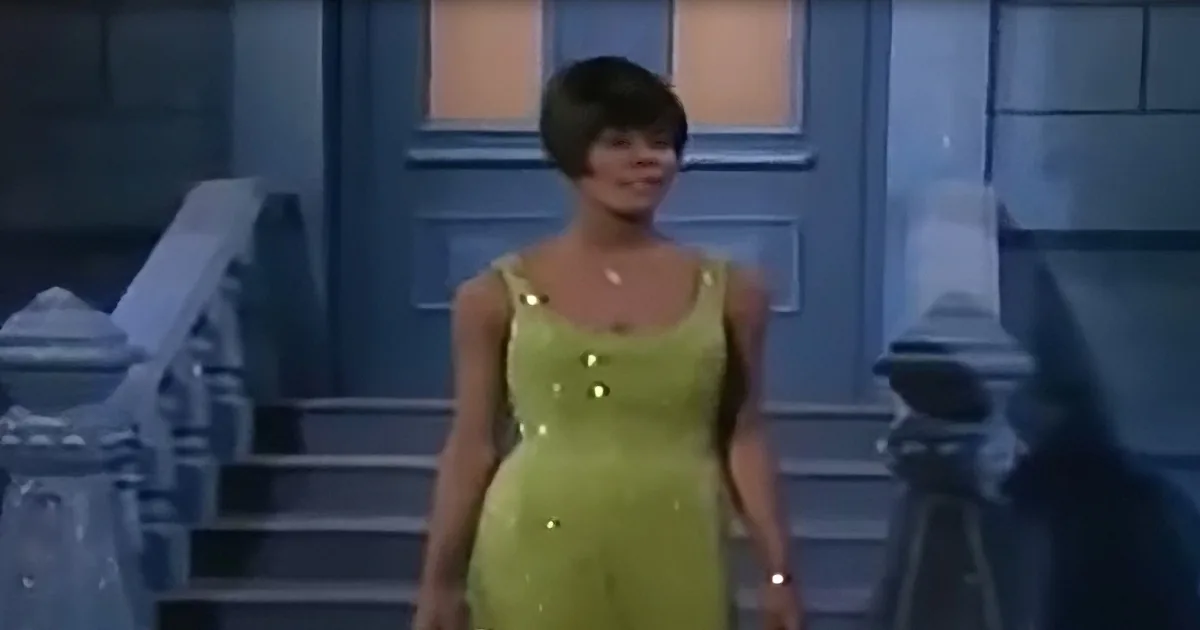 A woman in a bright green dress stands in front of a blue door, smiling.