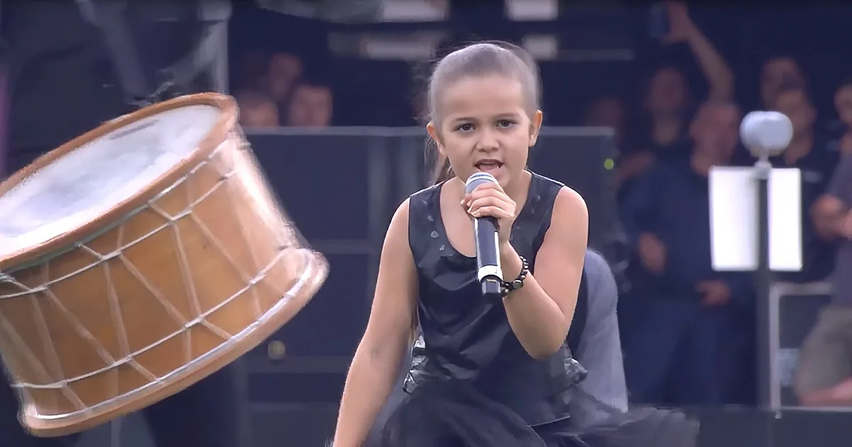 A young girl in a black dress sings into a microphone, with a large drum in the foreground and a blurred audience in the...