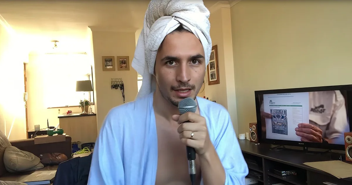 A man in a robe and towel on his head holds a microphone, speaking in a room with a TV and various items in the background.