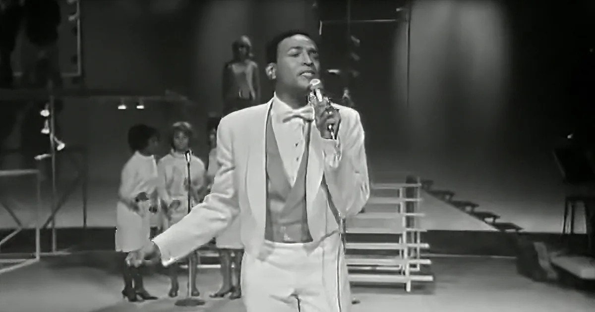 A man in a white suit sings into a microphone on stage, with backup dancers behind him.