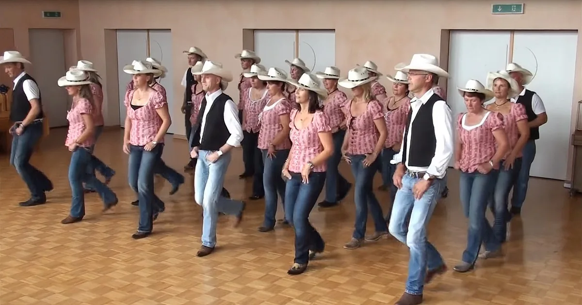 A group of people in cowboy attire is dancing in a room with wooden floors and white walls.