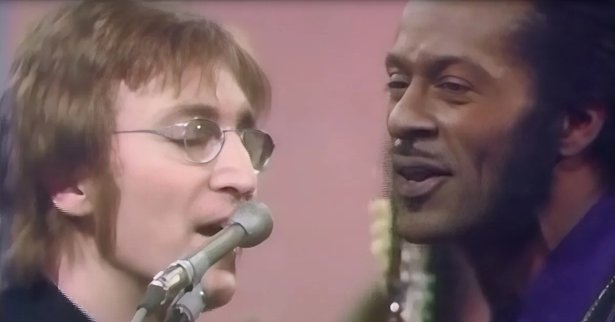 A man with glasses sings into a microphone, another man in a purple shirt smiles nearby.