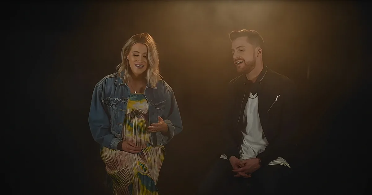 A pregnant woman in a denim jacket and colorful dress holds her belly, smiling, with a man in a black jacket sitting...
