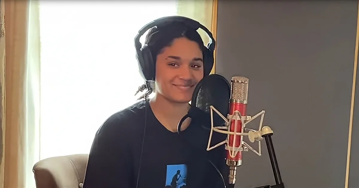A person wearing headphones sits in front of a microphone, smiling, in a recording studio setup.