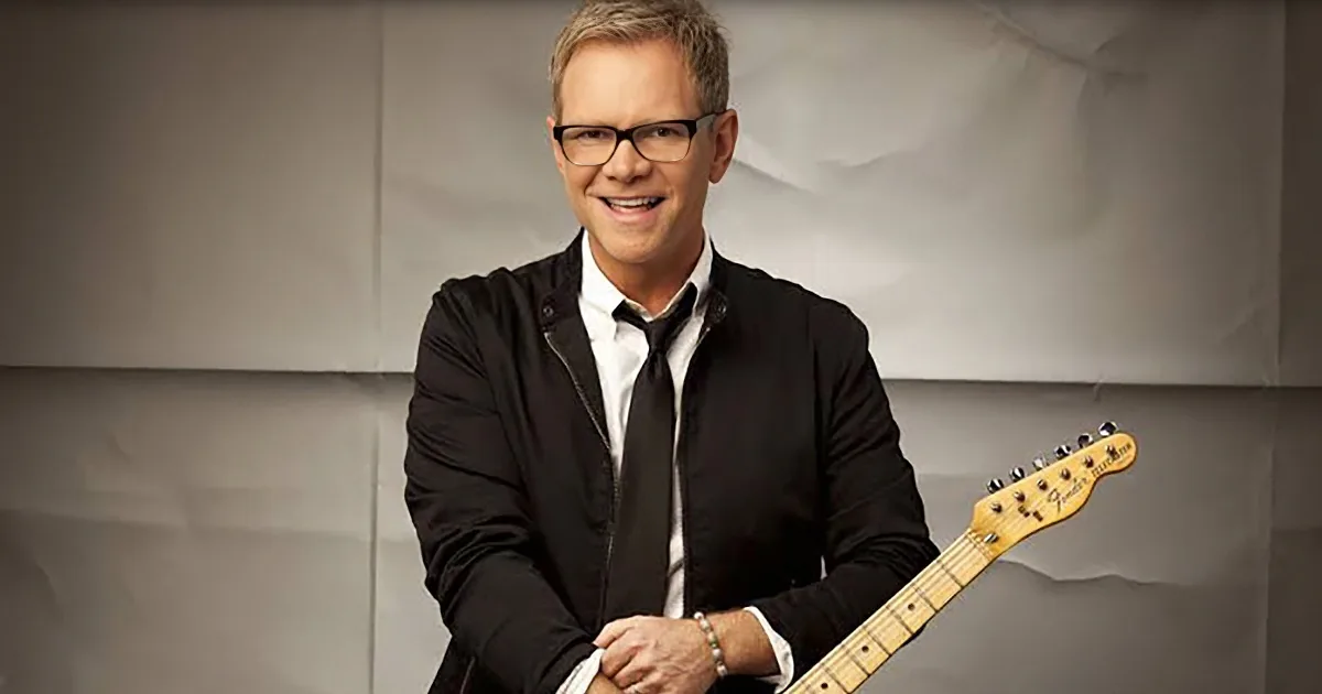 A man in glasses and a black jacket holds a Fender guitar, smiling against a textured background.