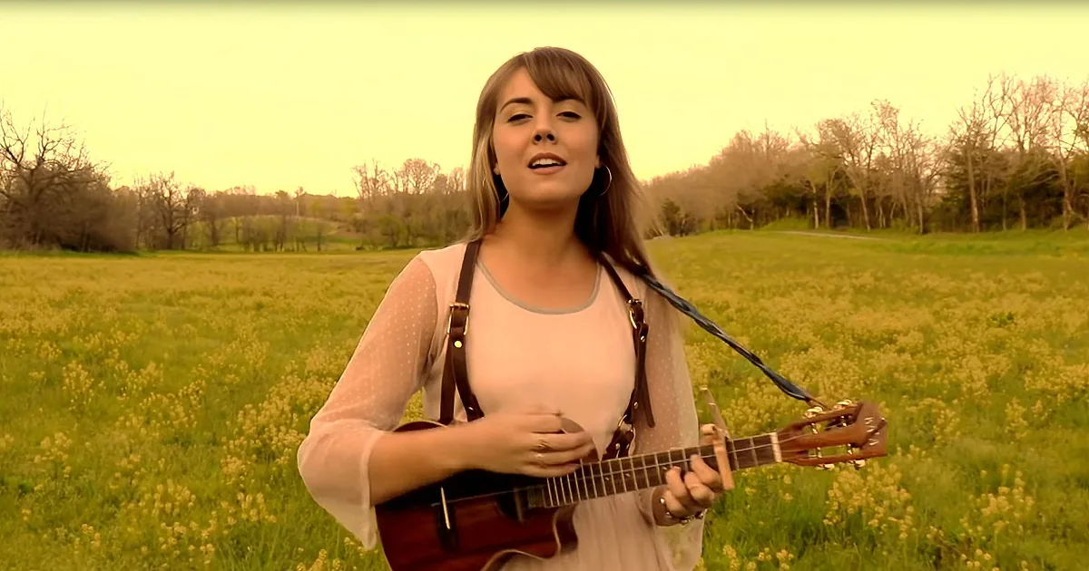 A woman in a field holds a guitar, singing with a serene backdrop of trees and grass.