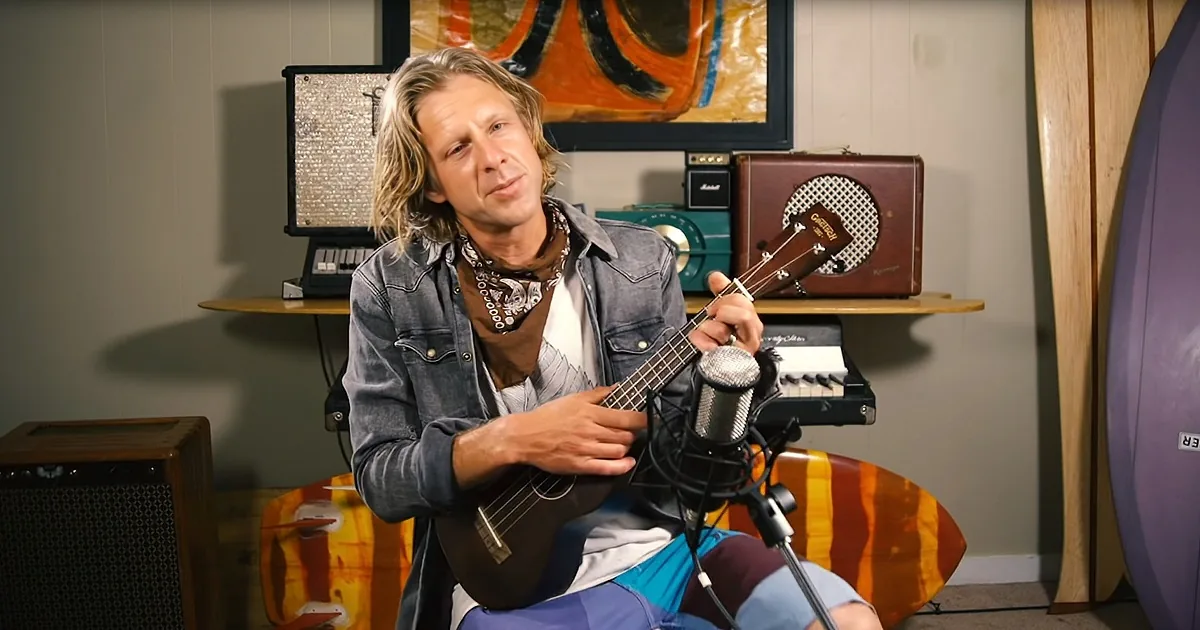 A man with long hair and a bandana plays a ukulele in a room with musical equipment and a surfboard.