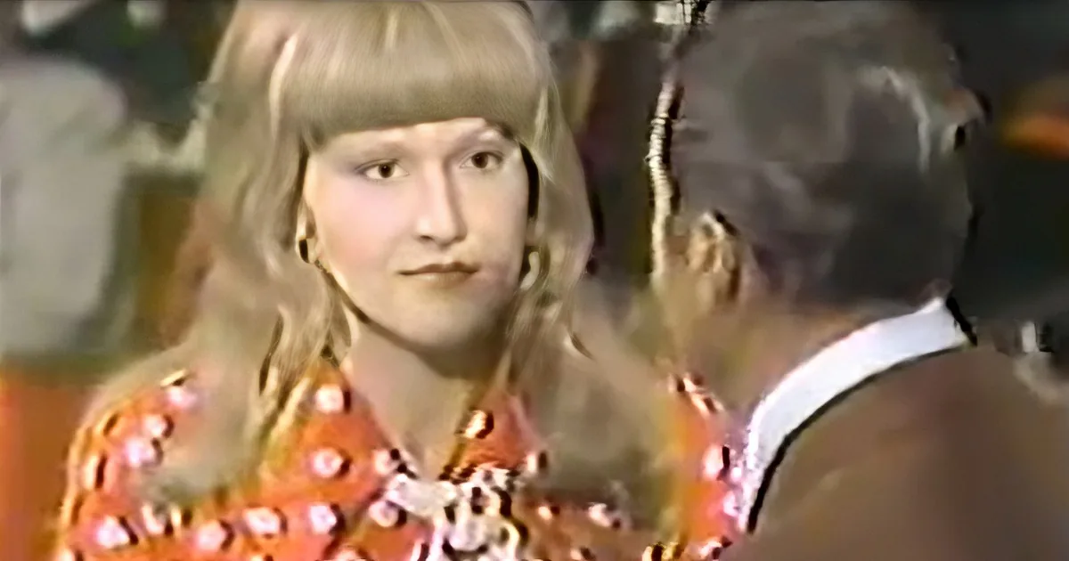 A woman with blonde hair and a patterned orange dress is looking at a man in a suit.