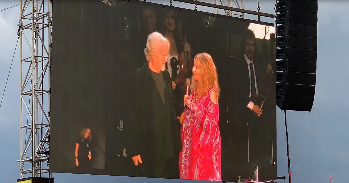 A woman in a red dress holds a microphone on stage, with a large screen showing a man in a suit and another person.