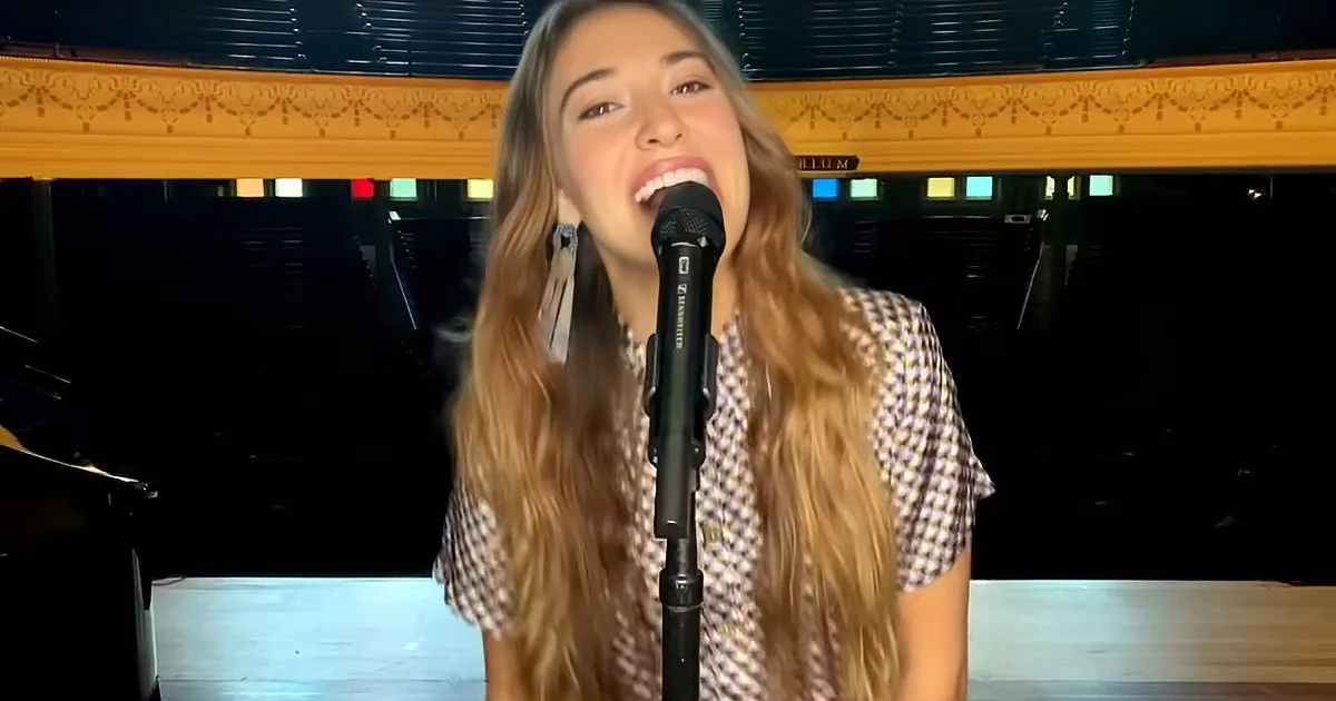 A person with long hair sings into a microphone in front of a piano and colorful background.