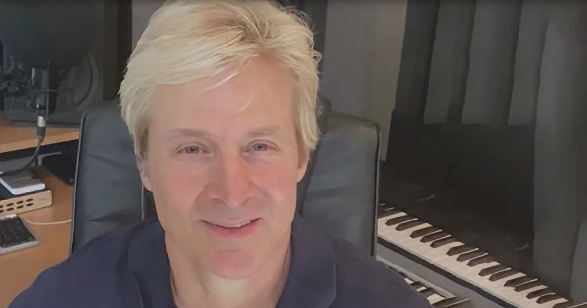 A person with blonde hair sits in a black chair, smiling, with a piano and desk in the background.