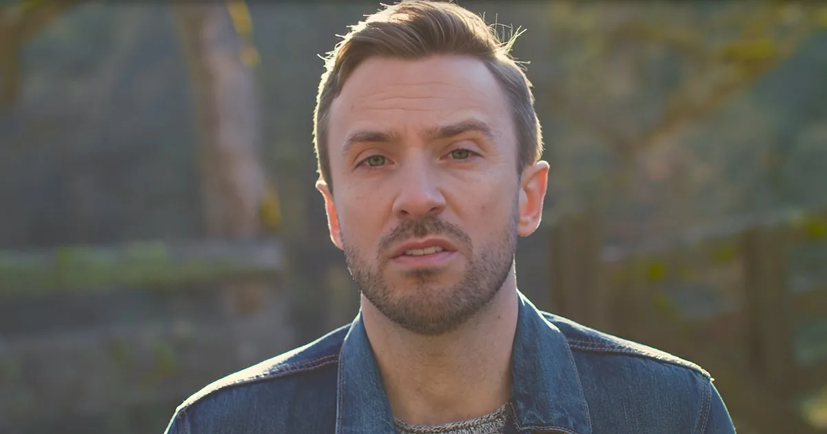 A man with short brown hair and a beard, wearing a denim jacket, stands outdoors with a blurred background of trees.