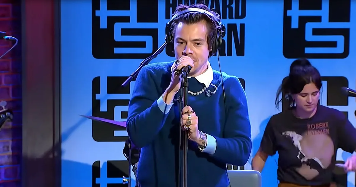 A man in a blue sweater sings into a microphone on stage, wearing headphones and a pearl necklace.