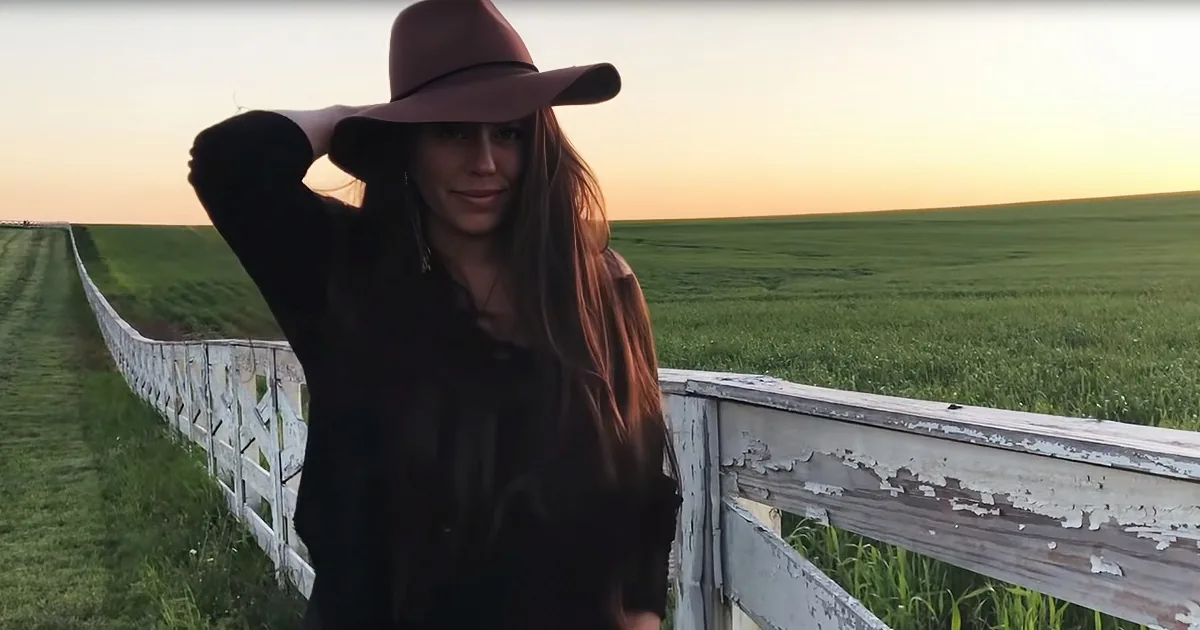 A person in a wide-brimmed hat stands near a weathered fence, with a vast green field and sunset in the background.