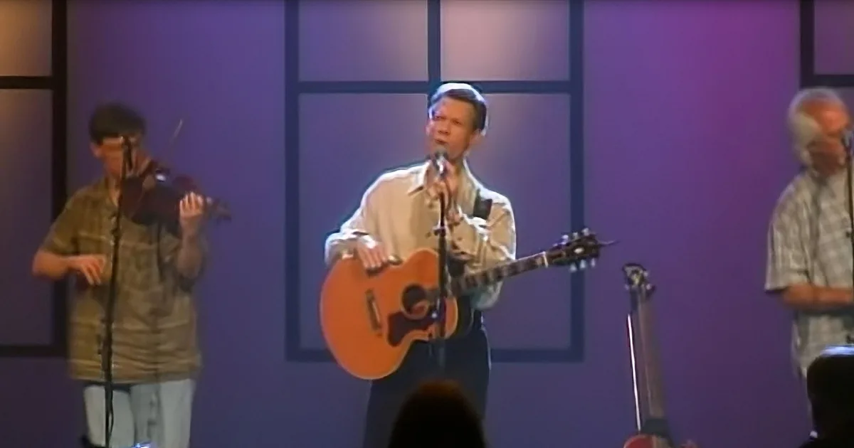 A man in a white shirt plays guitar and sings on stage, accompanied by musicians.