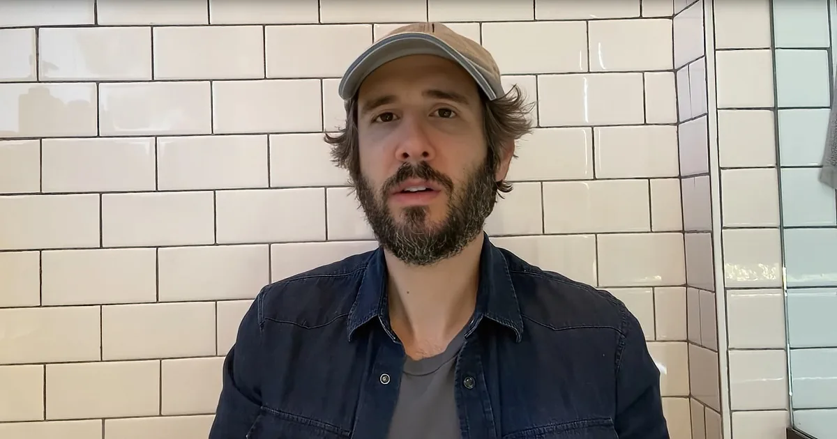 A man with a beard and cap stands in front of white subway tiles, wearing a denim shirt.