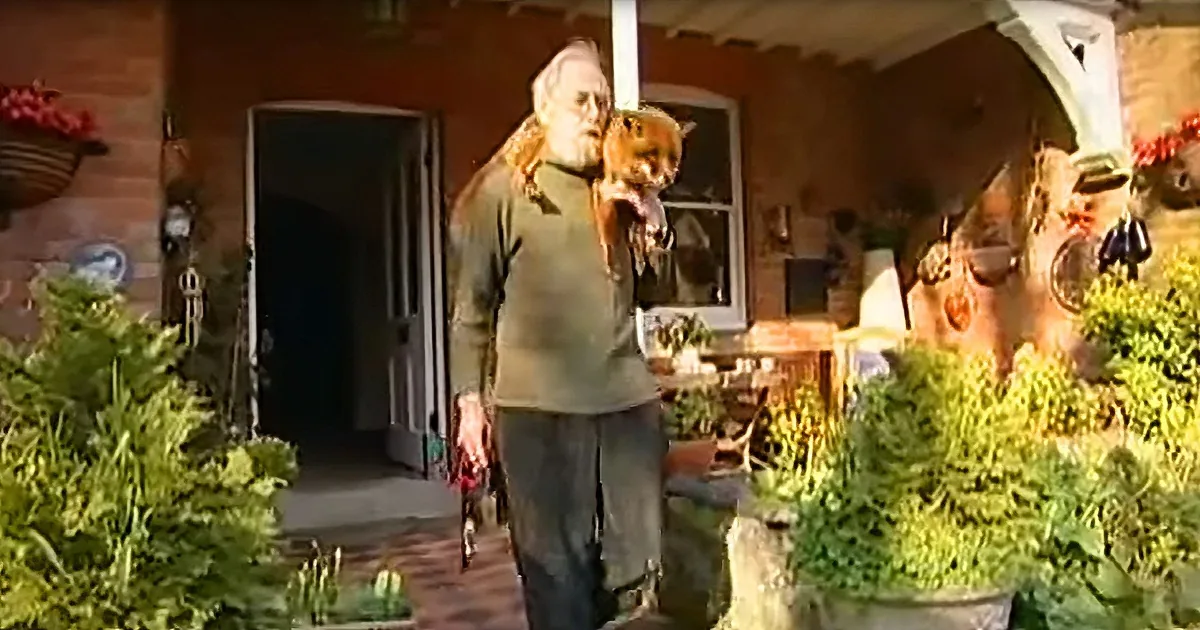 An elderly man holds a cat while walking on a porch, surrounded by greenery and a brick path.