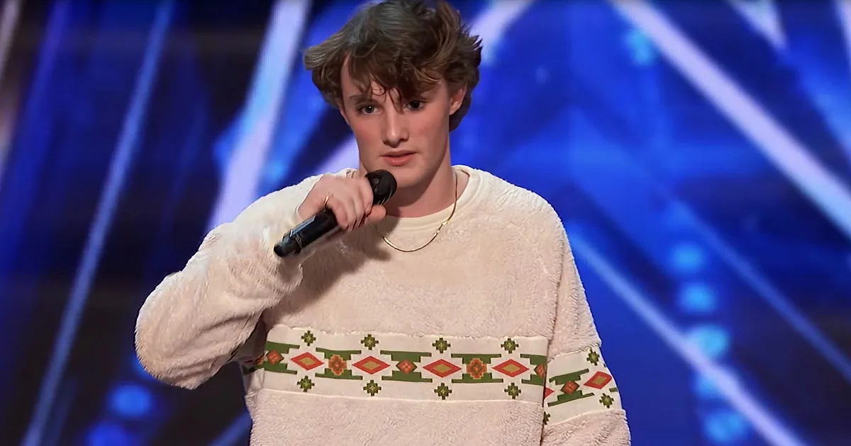 A person wearing a white sweater with colorful patterns is holding a microphone on stage.