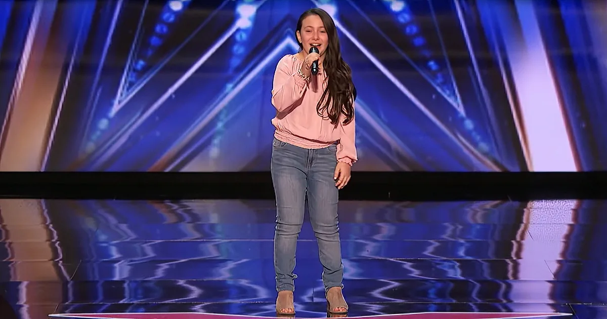 A girl in a pink sweater and jeans sings on stage with a microphone, surrounded by a colorful, illuminated backdrop.