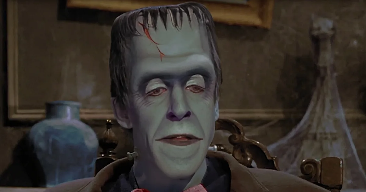A person with pale makeup and dark hair, resembling Frankenstein, sits in a room with ornate furniture and a vase.