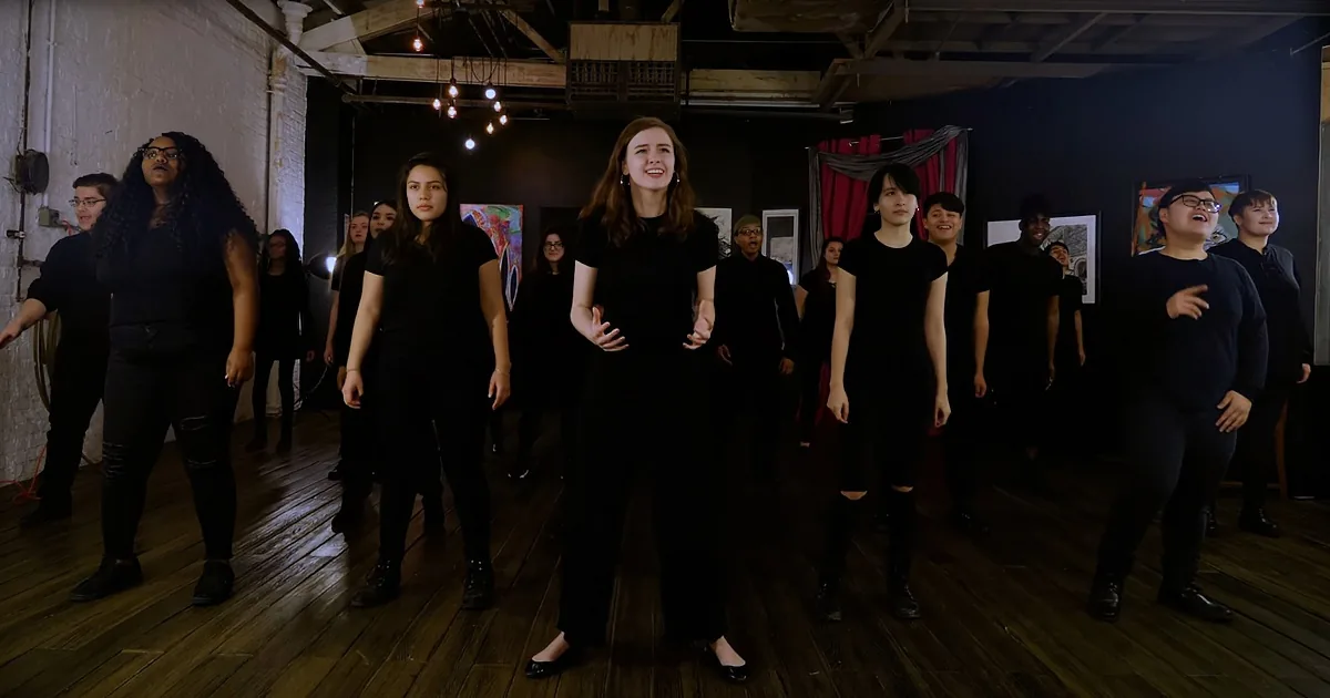 A woman in black leads a group of people in a dance routine in a dimly lit studio.