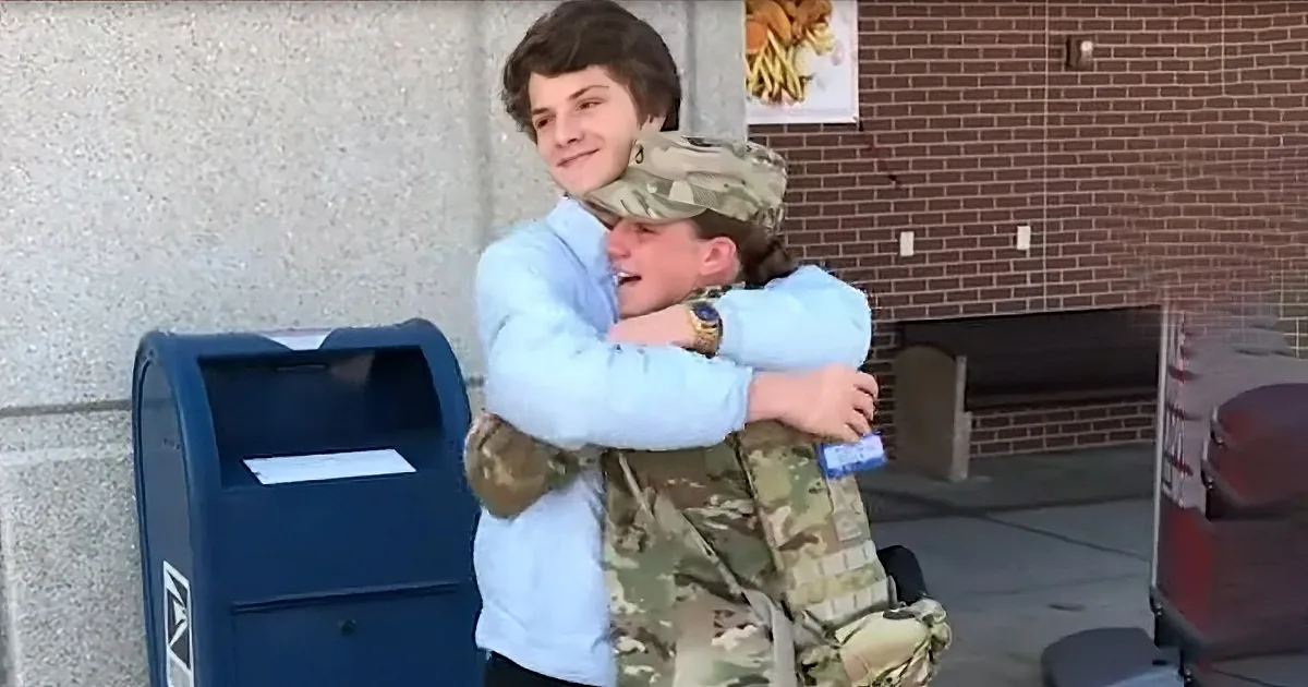 A soldier in camouflage embraces a young person in a light blue shirt near a blue mailbox.