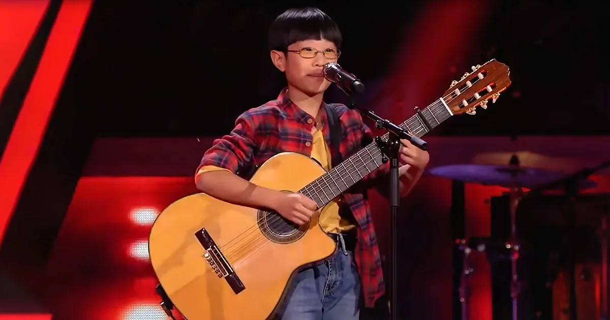 A young boy in a plaid shirt plays an acoustic guitar on stage with a microphone.