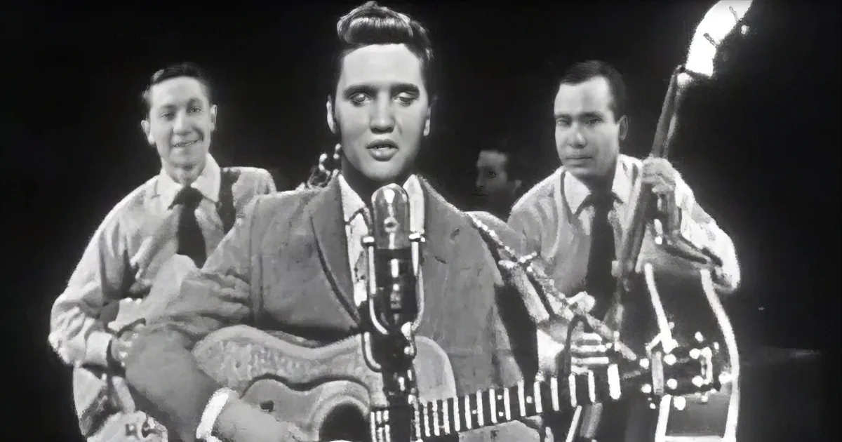 Elvis Presley sings into a microphone, holding a guitar, with two musicians behind him.