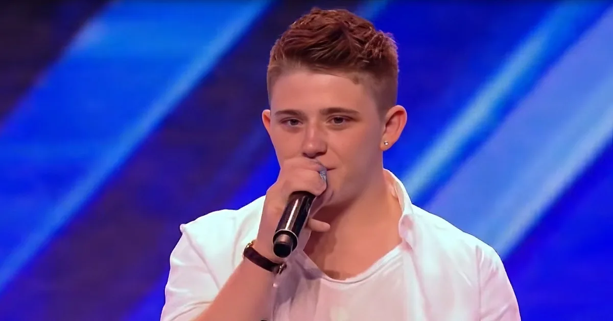 A young man with short brown hair, wearing a white shirt, sings into a microphone on a stage with blue lighting.