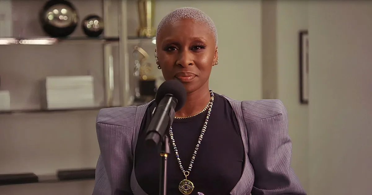 A woman with short hair, wearing a purple blazer and necklace, speaks into a microphone in a well-lit room.