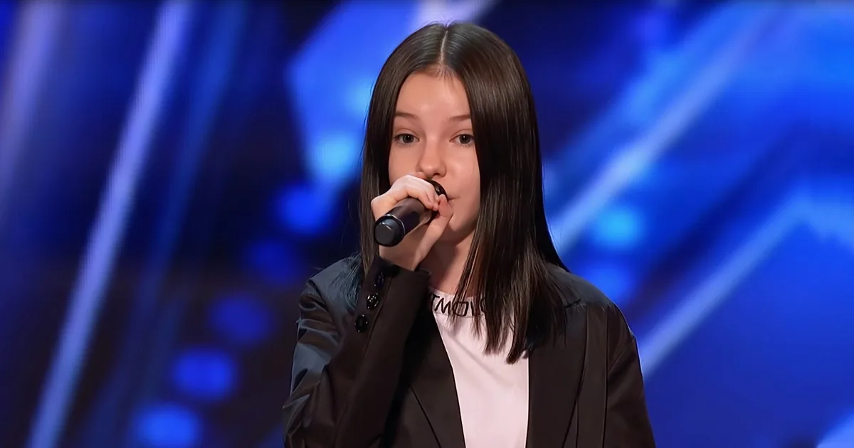 A person with long dark hair holds a microphone, wearing a black blazer over a white shirt, against a blue stage backdrop.