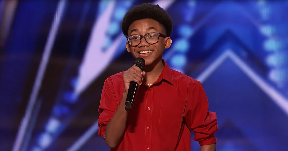 A person in a red shirt holds a microphone, smiling, with a blue stage background.