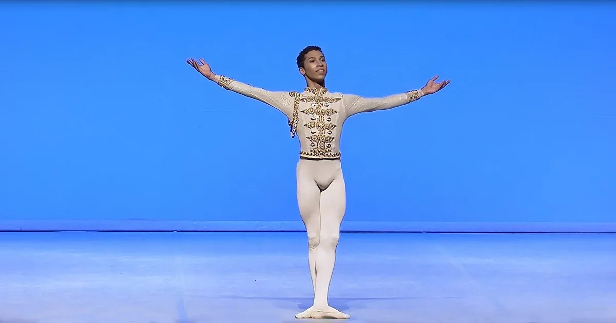 A ballet dancer in a white leotard with gold embellishments stands on pointe with arms outstretched.