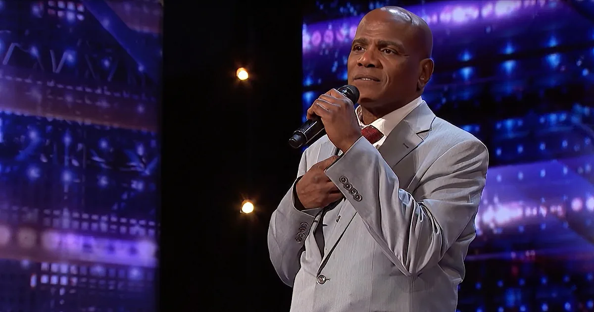 A man in a white suit holds a microphone, standing on a stage with a colorful backdrop.