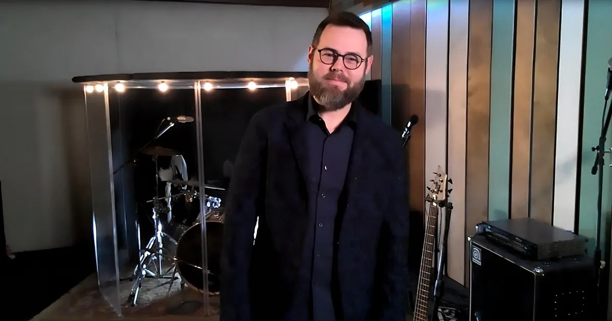 A man with a beard and glasses stands in a music studio with a drum set, guitar, and sound equipment.