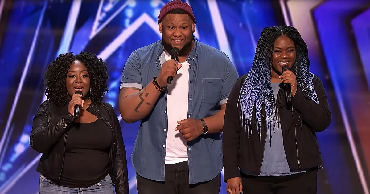 Three individuals stand on stage, each holding a microphone, with a vibrant blue and white background.