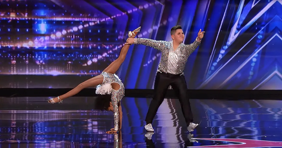 A dancer in a silver outfit performs a split while another dancer in a sequined top and black pants points dramatically.