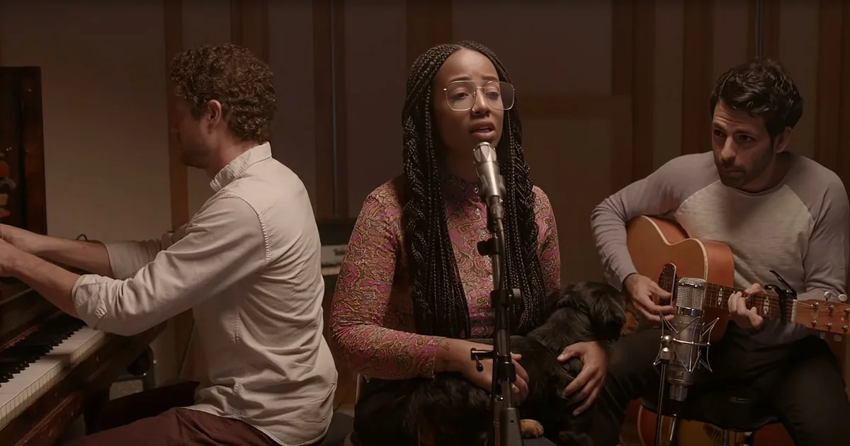 A woman sings into a microphone, accompanied by a pianist and guitarist in a recording studio.