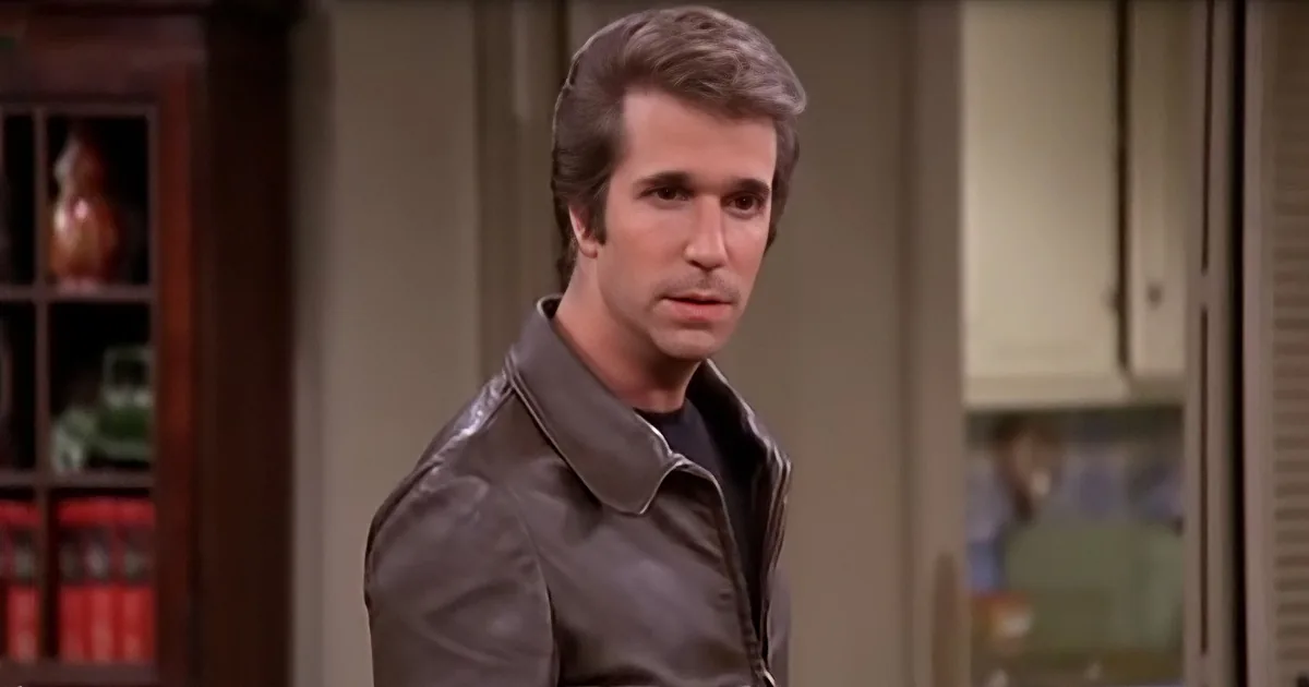A man in a brown leather jacket stands in a kitchen, looking to the side with a serious expression.