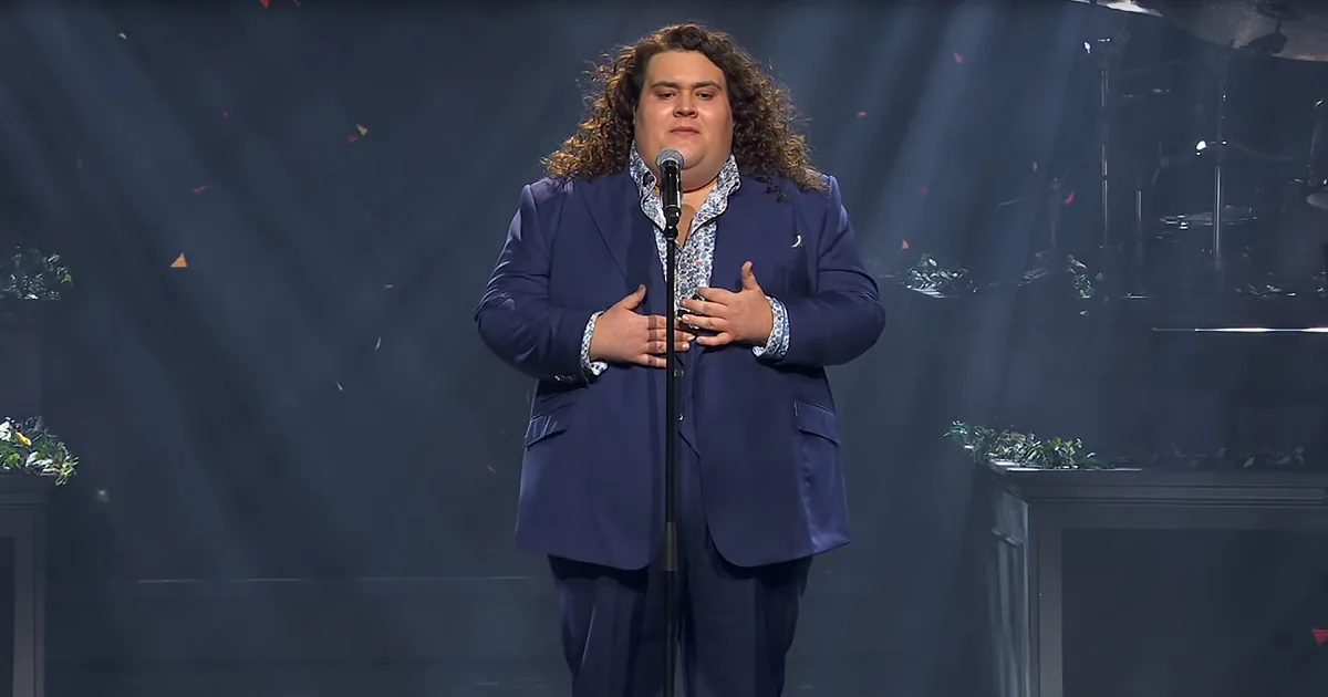 A person in a blue suit stands on stage with hands on chest, microphone in front, surrounded by stage lights and confetti.