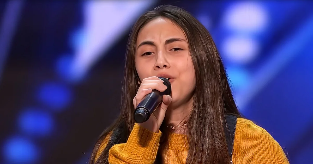 A person in a yellow sweater sings into a microphone, with a blurred blue background.